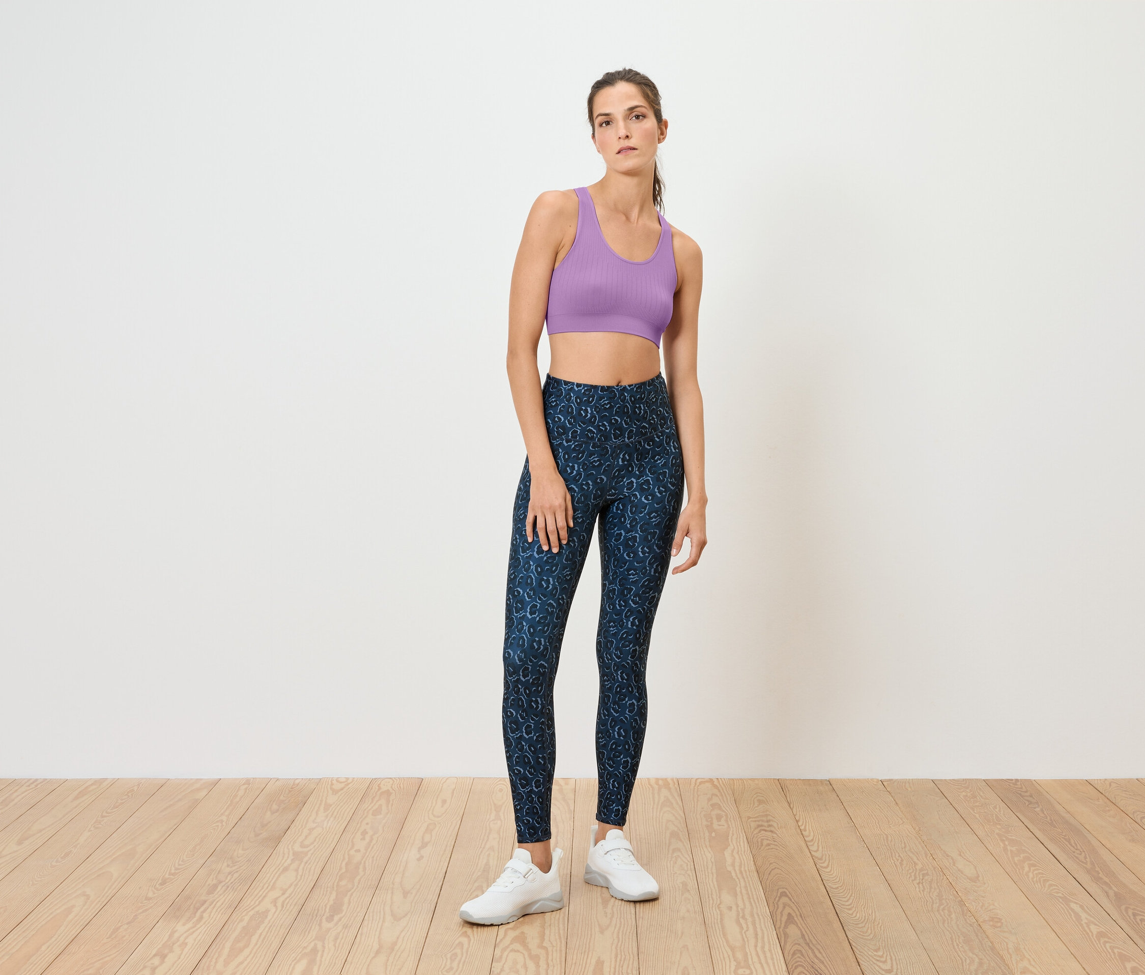 Une femme se tient sur un plancher en bois, portant un soutien-gorge de sport violet et un legging bleu.