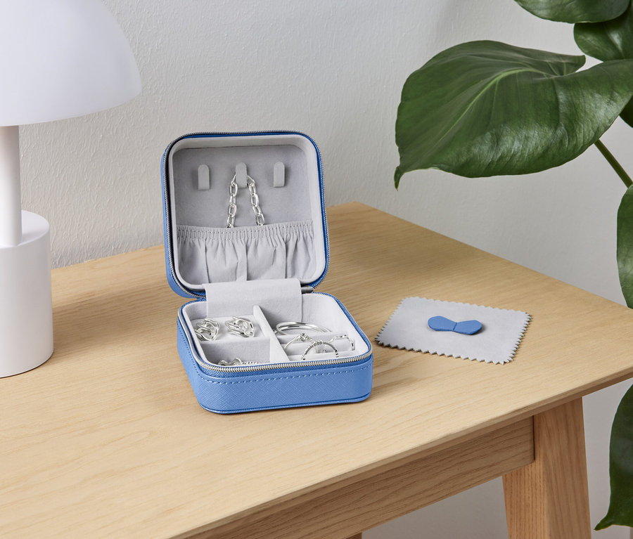 Une boîte à bijoux bleue ouverte avec des bijoux en argent est posée sur une table en bois à côté d'une lampe blanche et d'une plante verte.