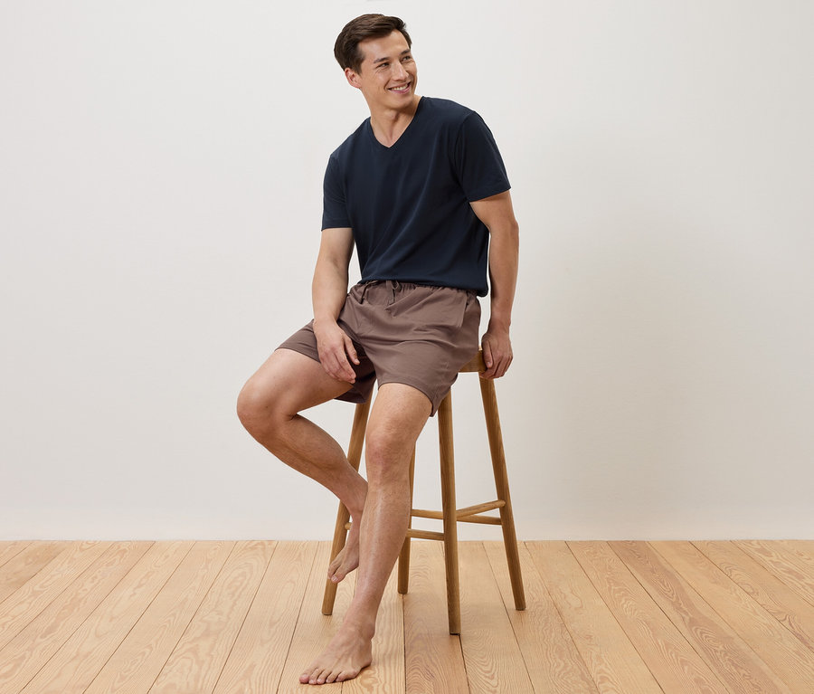 Un homme est assis pieds nus sur un tabouret, portant un t-shirt bleu marine et un short marron.