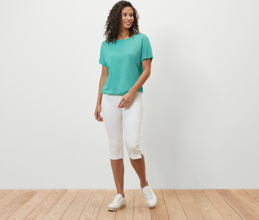 Une femme pose en chemisier turquoise à encolure dégagée et pantalon capri blanc.