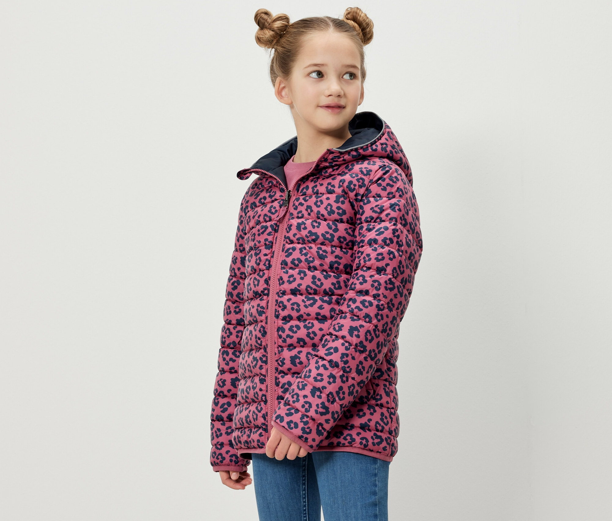 Une fille pose en veste softshell et matelassée 2 en 1 pour enfants. La veste a un fond rose avec un motif animal bleu marine.