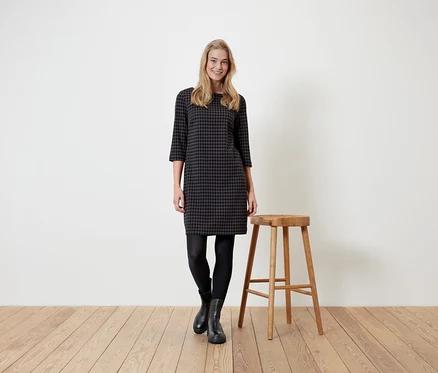 Une femme se tient à côté d'un tabouret en bois et porte une robe pied-de-poule, des leggings noirs et des bottes.