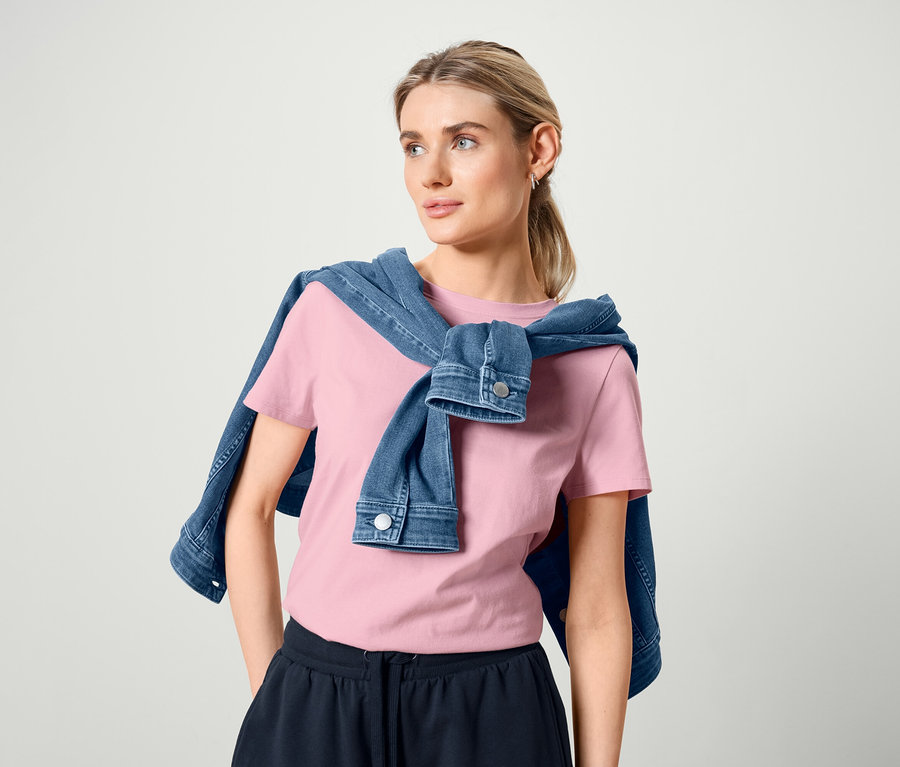 Femme avec veste en jean sur les épaules, portant un t-shirt basique rose et un sweatshort bermuda.