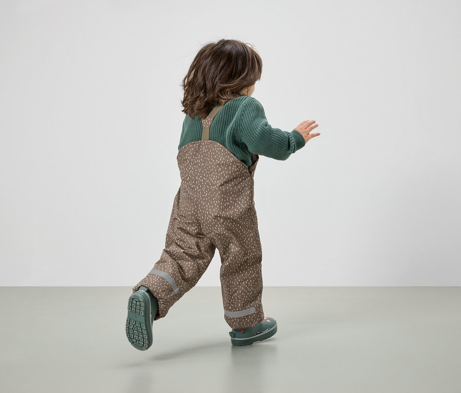 Un enfant en salopette marron et pull vert court en s'éloignant du spectateur.