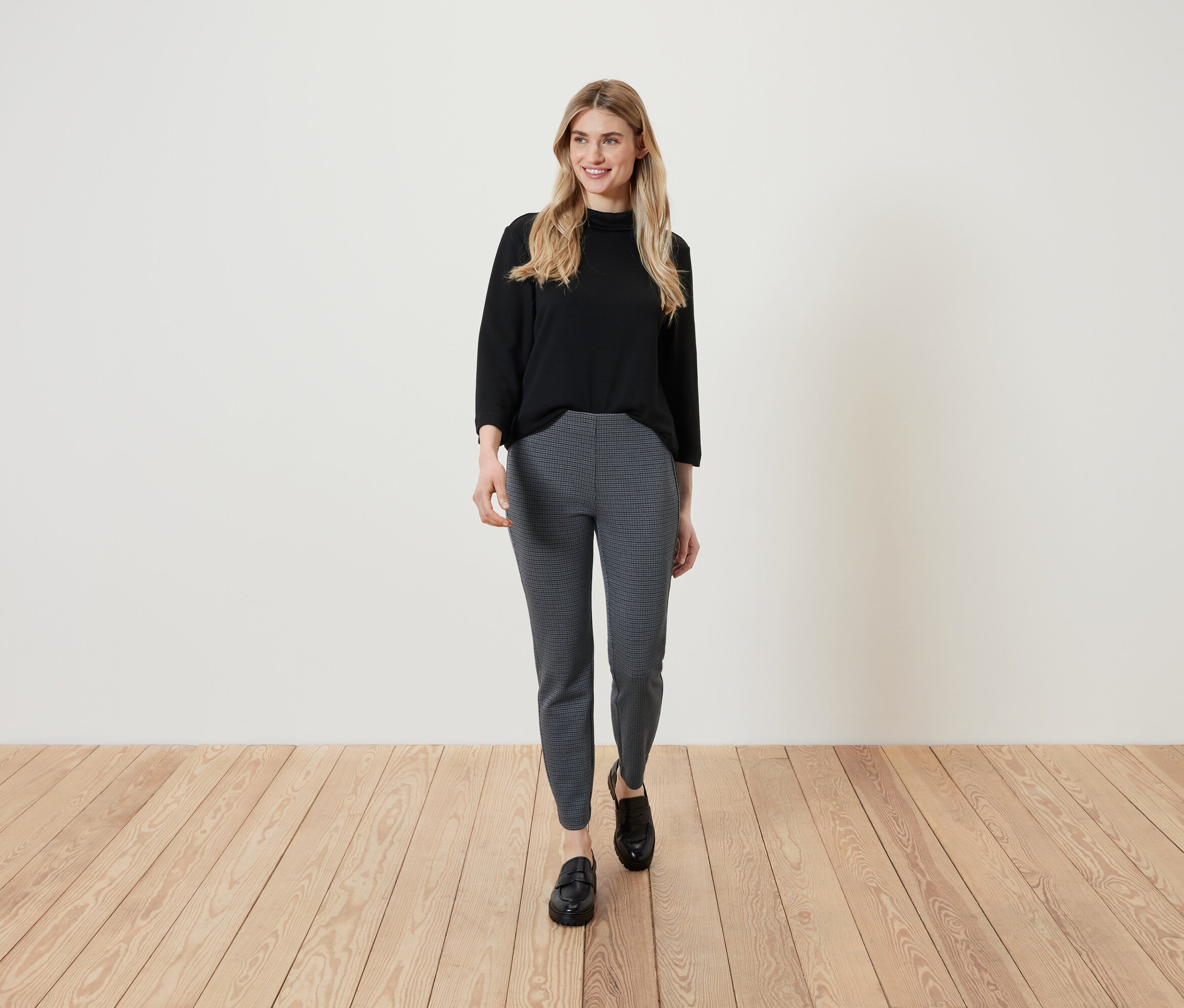 Une femme blonde portant un haut noir et un pantalon à motifs marche sur un plancher en bois.