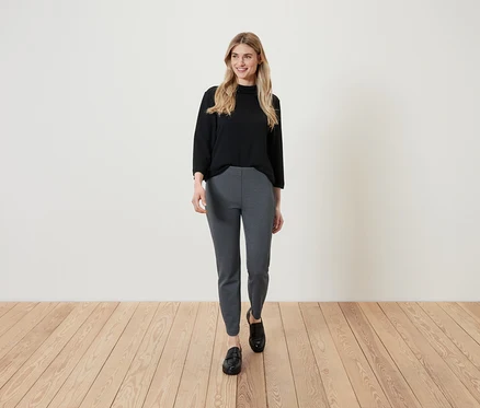 Une femme blonde portant un haut noir et un pantalon à motifs marche sur un plancher en bois.