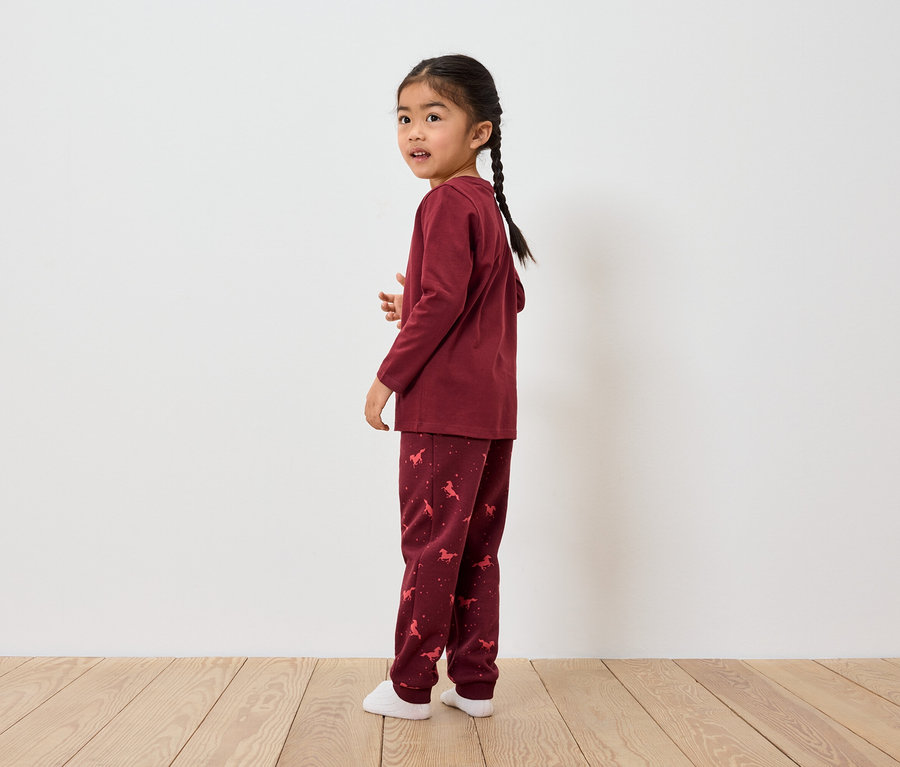Une fillette asiatique avec une tresse se tient de côté et regarde par-dessus son épaule. Elle porte un pyjama bordeaux avec un motif de cheval.