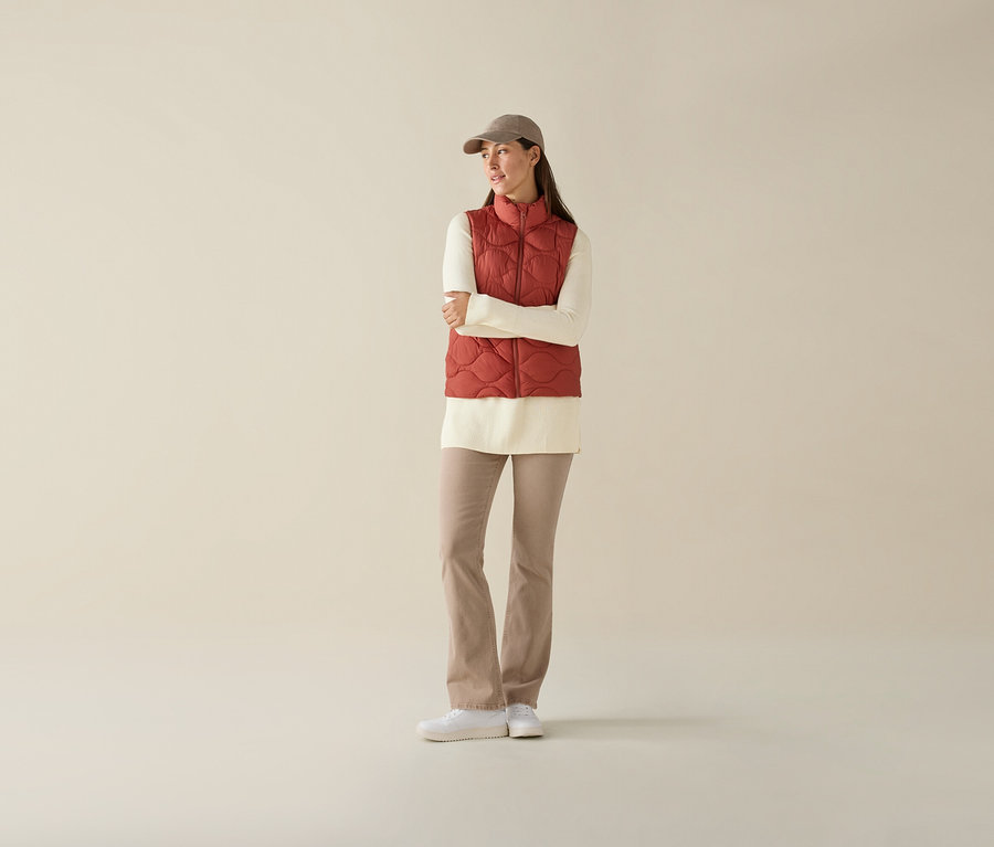 Une femme les bras croisés porte un gilet matelassé sans manches, un haut couleur crème, un pantalon beige et une casquette.