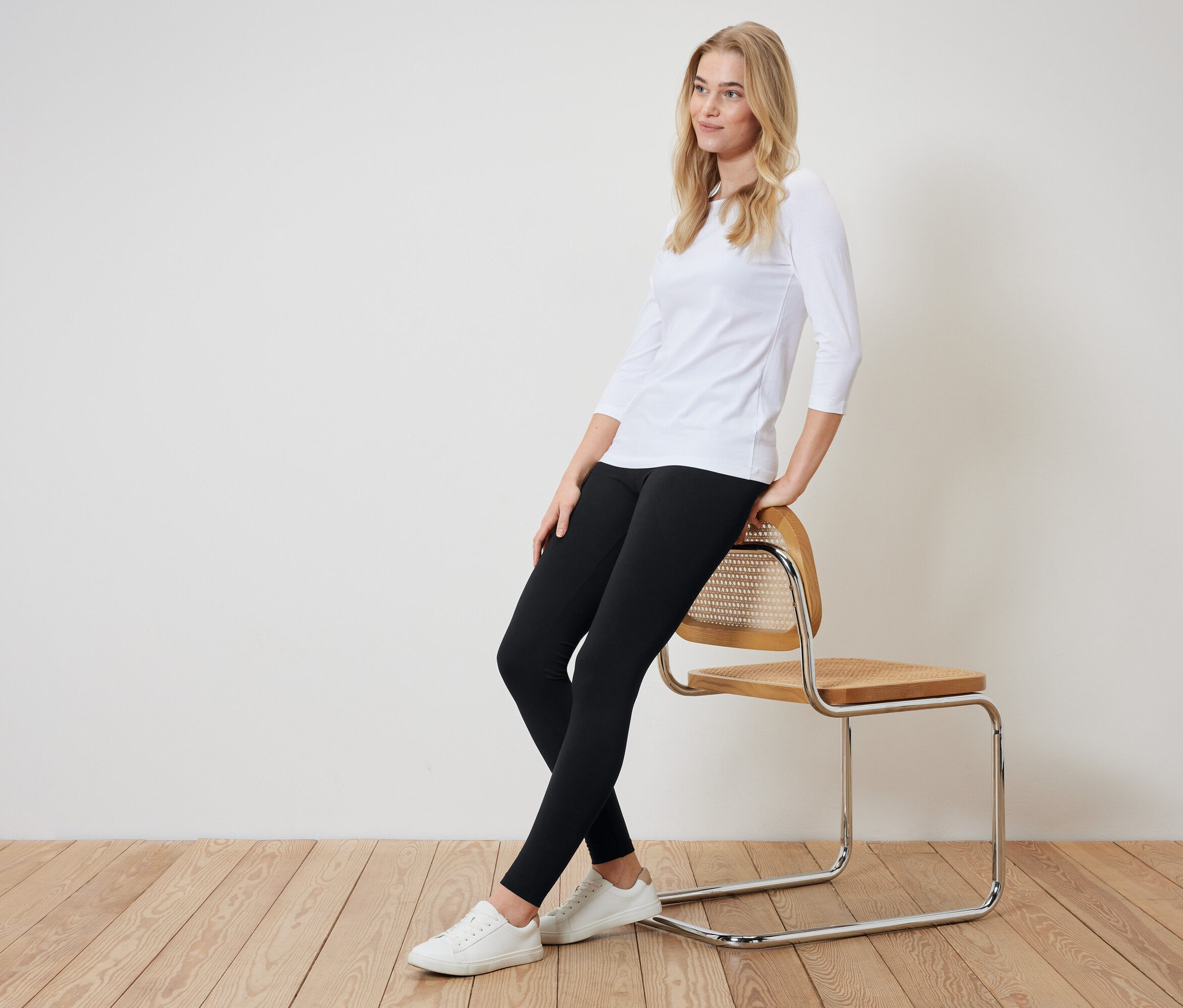 Une femme blonde est assise de côté sur une chaise. Elle porte un t-shirt blanc à manches trois-quarts, un legging noir et des baskets blanches.
