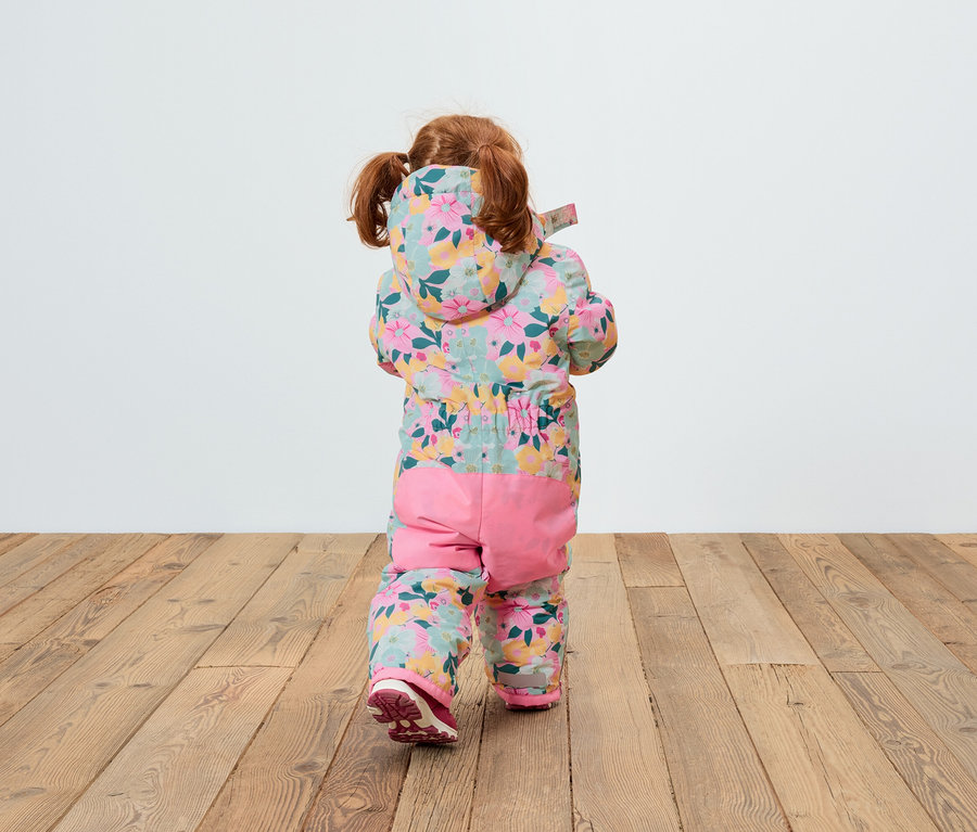 Vue arrière d'un enfant aux cheveux roux portant une combinaison à capuche fleurie et un pantalon rose, debout sur un plancher en bois.