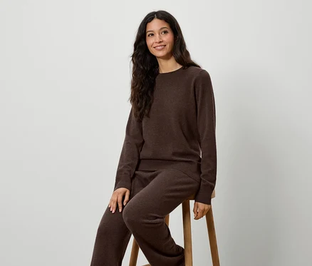 Une femme est assise sur un tabouret et porte un pull et un pantalon en tricot marron.