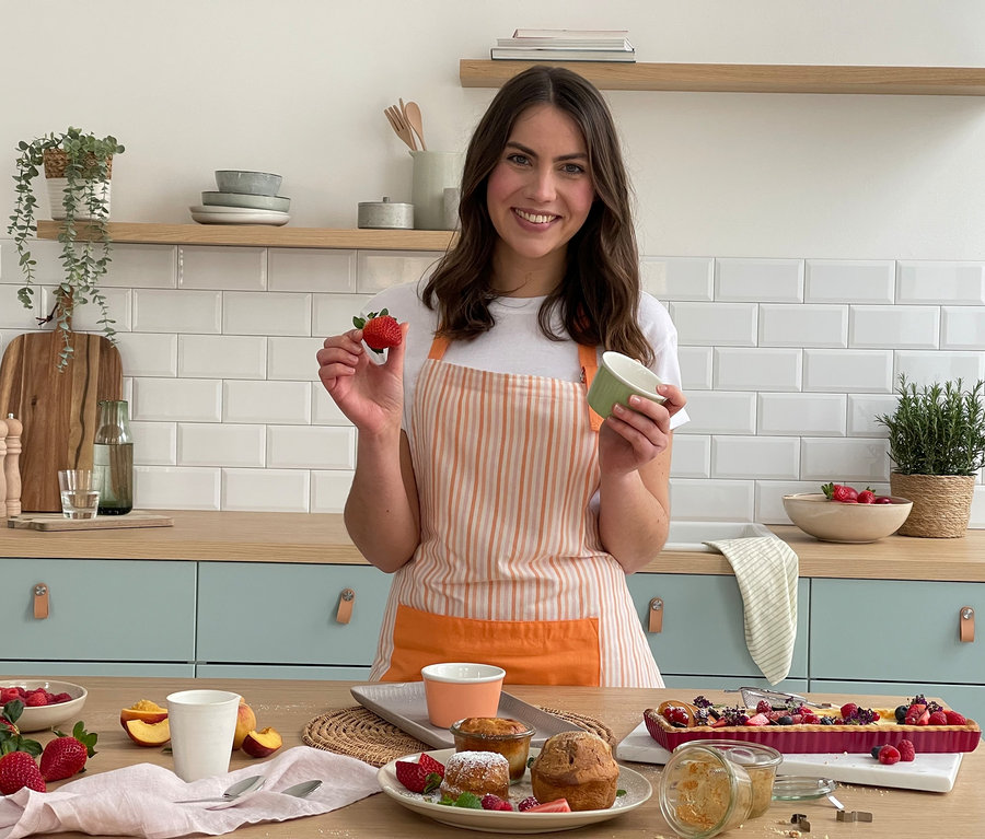 Une femme tient une fraise et un bol vert dans une cuisine. Sur la table, il y a 4 coupes à glace, des muffins et une tarte aux fruits.