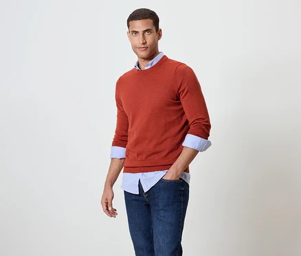 Un homme pose en pull orange sur une chemise bleue et un jean.