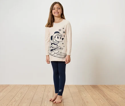 Une fille vêtue d'une tunique beige avec un imprimé de Mickey Mouse sur des skis et d'un legging bleu se tient sur un plancher en bois.