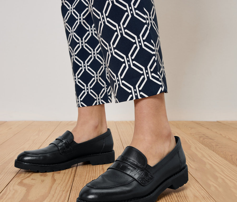 Jambes d'une personne portant des mocassins noirs et un pantalon bleu à motifs, debout sur un plancher en bois.