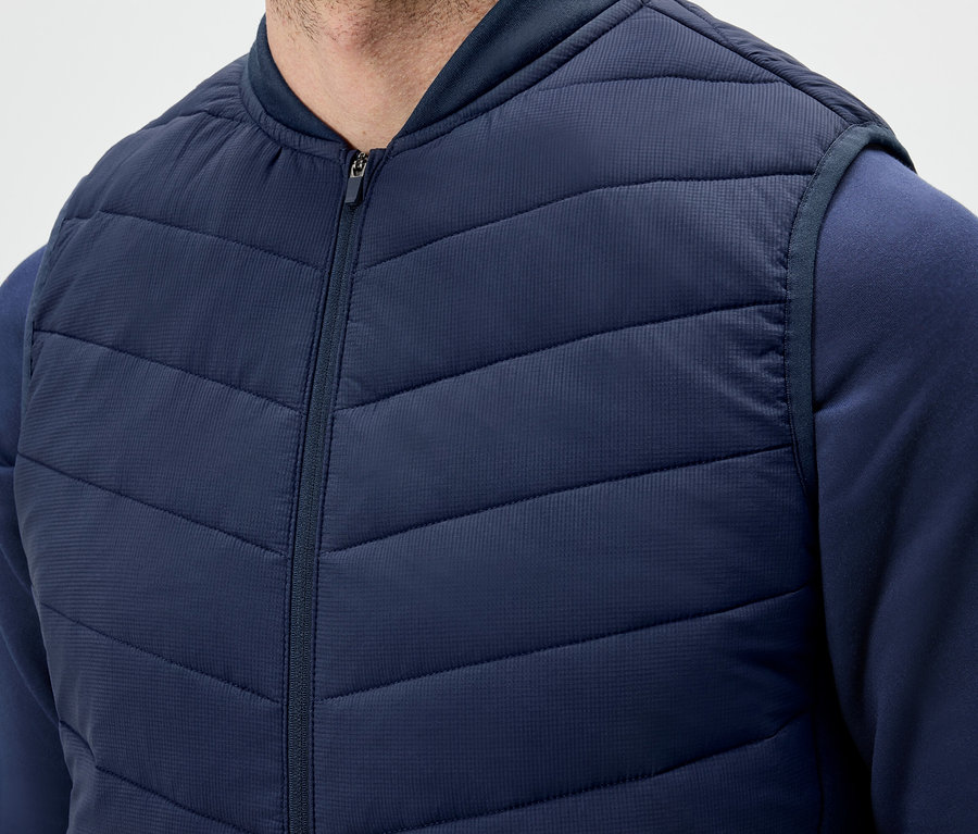 Un homme porte un gilet matelassé bleu foncé zippé et un sweat-shirt.
