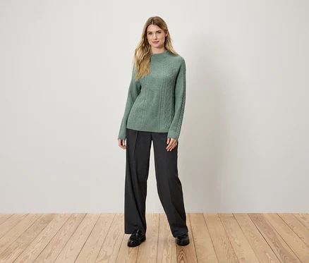 Une femme blonde se tient sur un plancher en bois et porte un pull en tricot vert et un pantalon noir.
