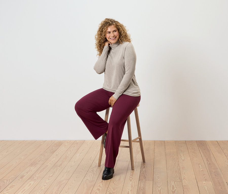 Une femme aux cheveux bouclés est assise sur un tabouret en bois, elle porte un pull à col roulé beige et un pantalon bordeaux.