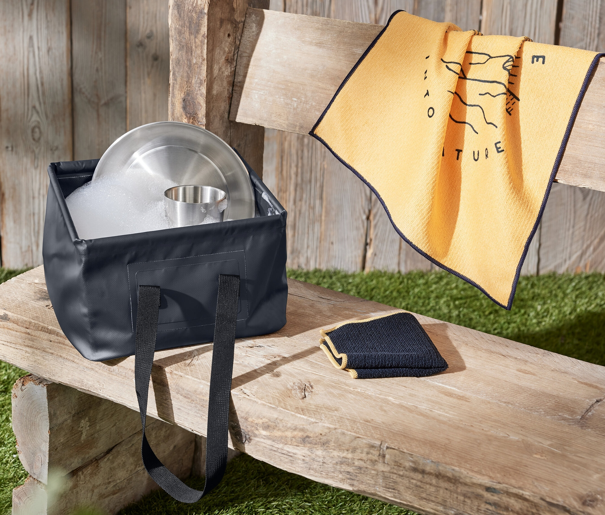 Un évier pliable rempli d'eau et de vaisselle est posé sur un banc en bois à côté d'une serviette.