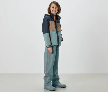 Un garçon portant une veste et un pantalon de pluie pour enfant, sauge, debout dans des bottes de pluie.