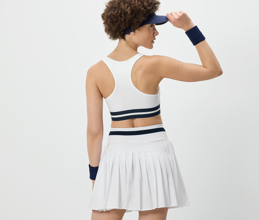 Femme vue de dos ajustant sa visière, portant un bustier de sport « Crop Top » blanc et une jupe plissée blanche.