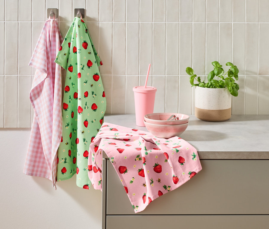 Trois torchons, une tasse rose avec une paille, deux bols roses et un pot de basilic sont posés sur un comptoir de cuisine.