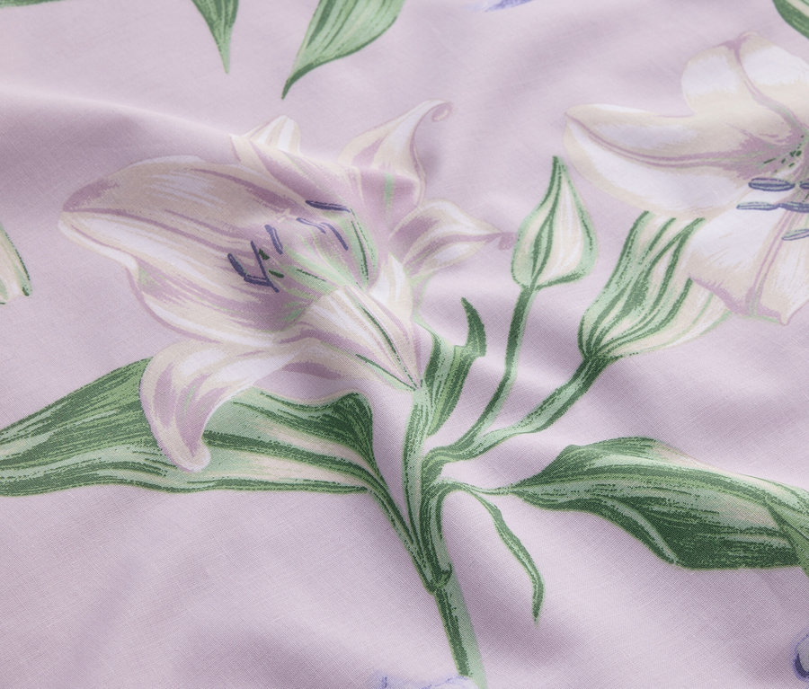 Gros plan d'un tissu avec un fond violet pâle, imprimé de lis blancs et de feuilles vertes.