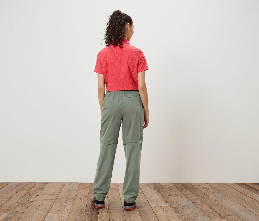 Vue arrière d'une femme aux cheveux bouclés portant une chemise rouge et un pantalon zippé vert olive sur un fond blanc.
