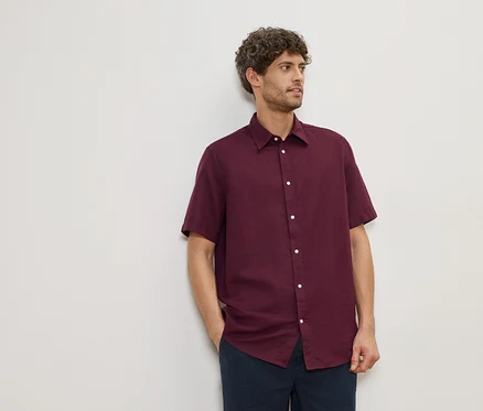 Un homme aux cheveux bouclés porte une chemise bordeaux à manches courtes et un pantalon bleu marine devant un mur blanc.