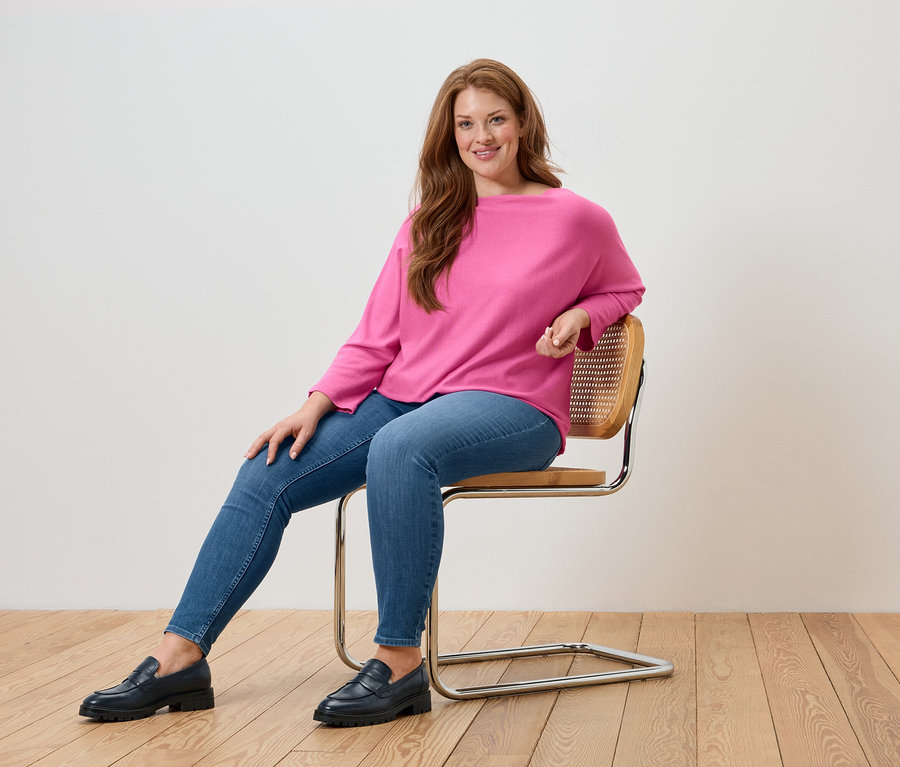 Une femme est assise sur une chaise et porte un pull rose, un jean bleu et des mocassins noirs.