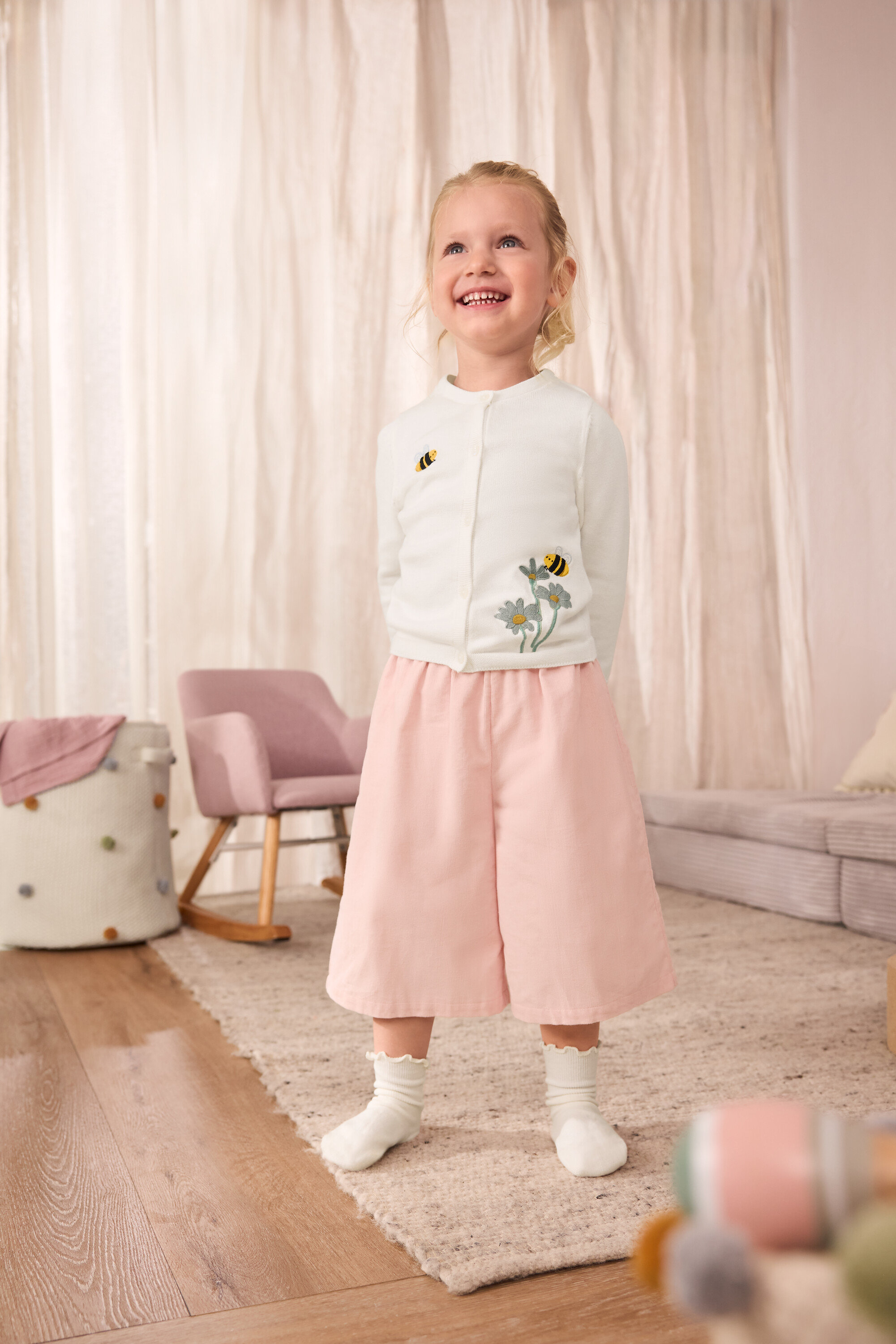 Une fillette souriante porte un gilet en tricot pour enfant avec des abeilles brodées, une culotte en velours côtelé pour enfant et 5 paires de chaussettes pour enfants à volants.