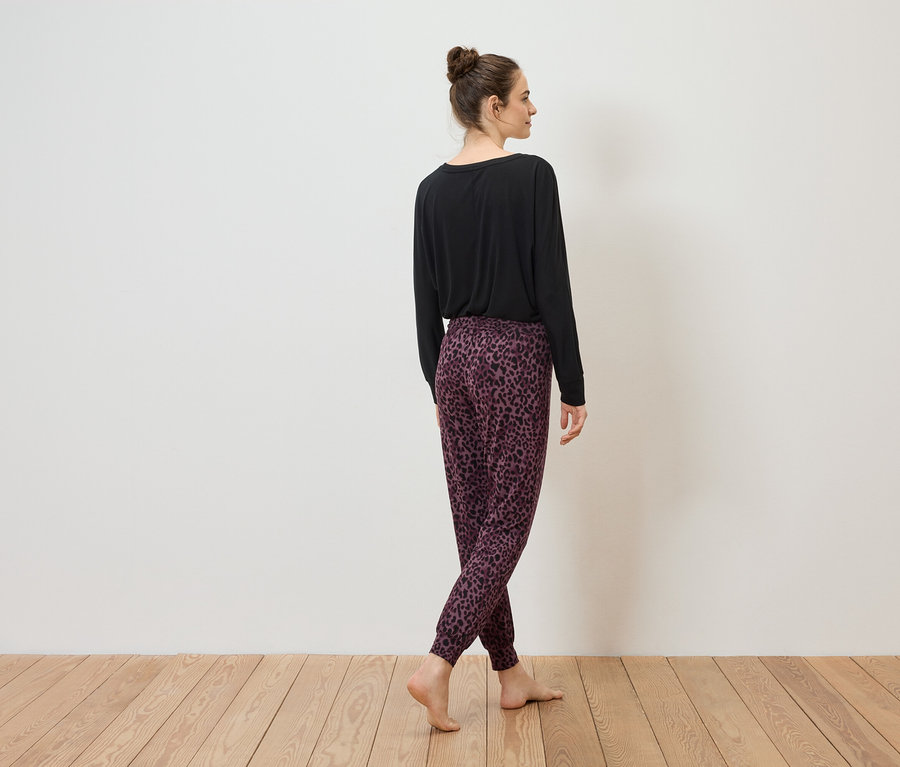 Femme vue de dos portant un haut noir et un pantalon de jogging violet et noir sur un plancher en bois devant un mur blanc.
