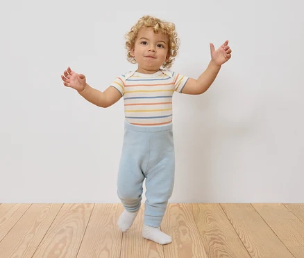 Un jeune enfant aux cheveux bouclés se tient debout, les bras écartés, portant un body rayé et un legging bleu clair.
