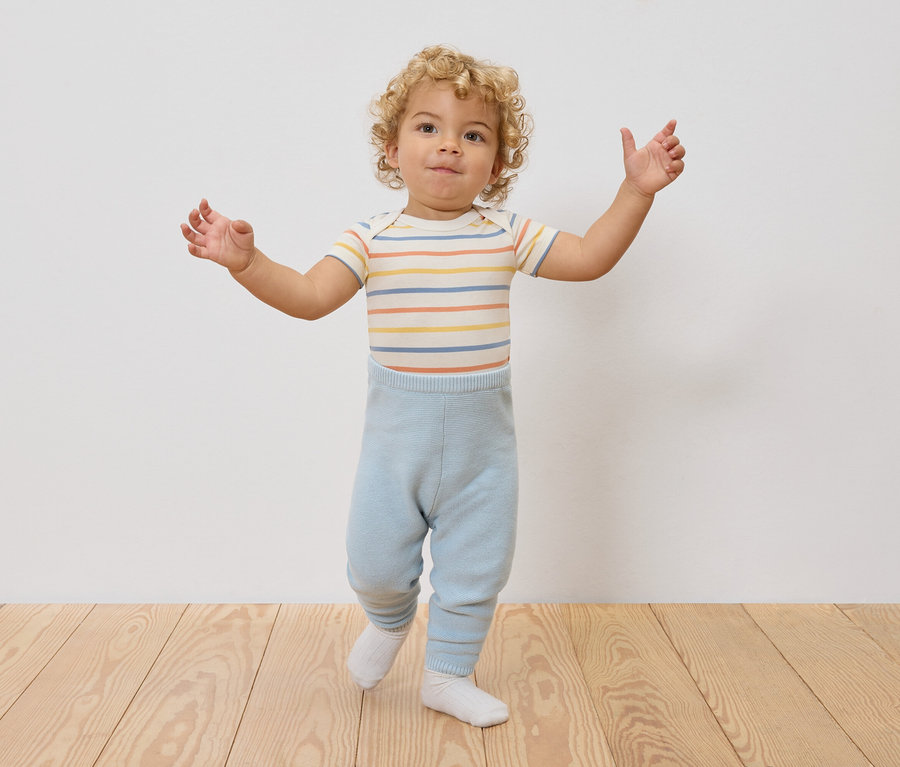 Un jeune enfant aux cheveux bouclés se tient debout, les bras écartés, portant un body rayé et un legging bleu clair.