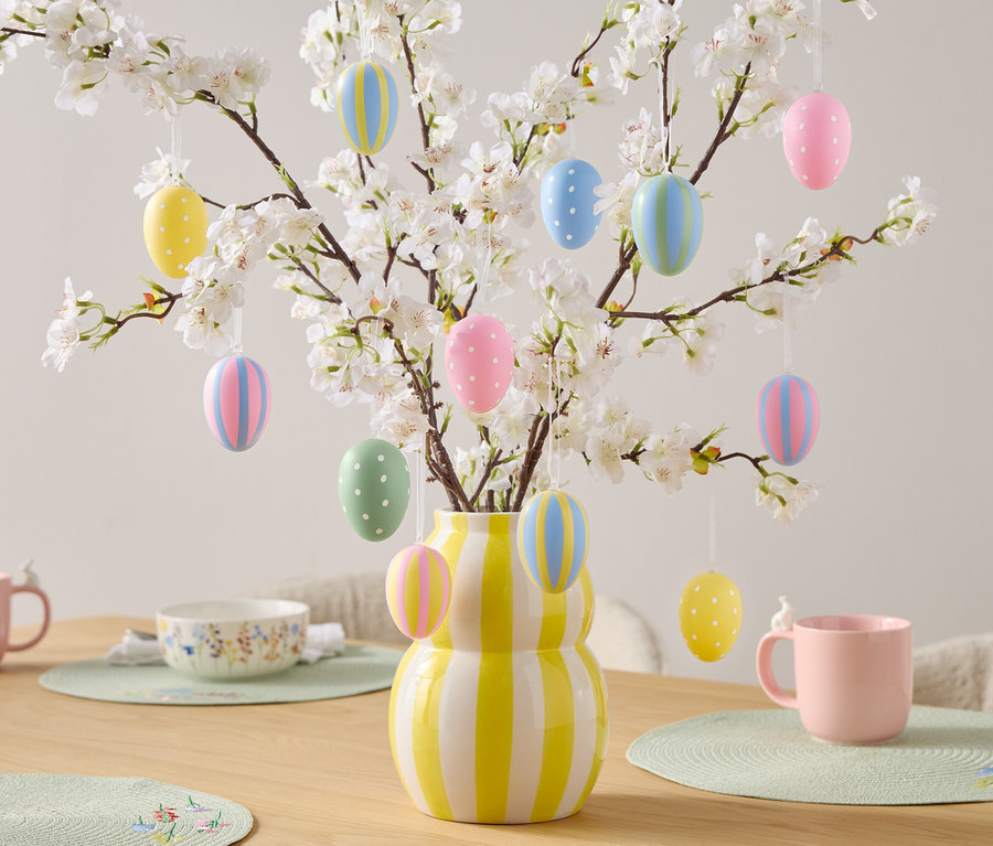 Table décorative avec œufs de Pâques sur des branches fleuries dans un vase rayé jaune et blanc, des tasses roses et un bol.