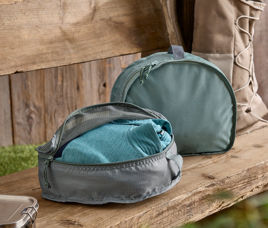 Cube de rangement gris avec un t-shirt bleu partiellement ouvert sur un banc en bois. À l'arrière-plan, un cube de rangement vert et un sac à dos.