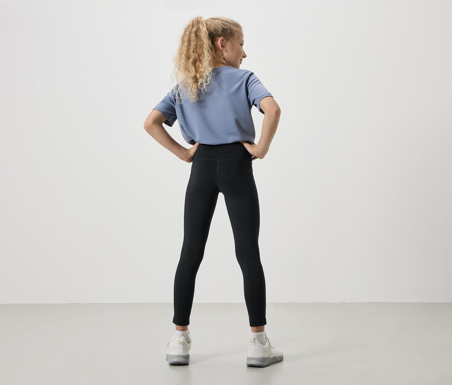 Vue arrière d'une jeune fille aux cheveux blonds bouclés, portant un t-shirt bleu, un legging noir et des baskets blanches, les mains sur les hanches.