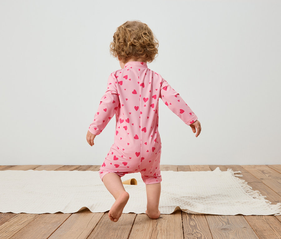 Un jeune enfant vu de dos, portant une combinaison rose avec un motif de cœurs, debout sur un tapis.