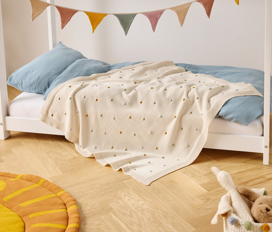 Un lit d'enfant avec des coussins bleus et une couverture crème à pois colorés se trouve sur un plancher en bois. Une bannière de fanions colorés est suspendue au-dessus du lit.