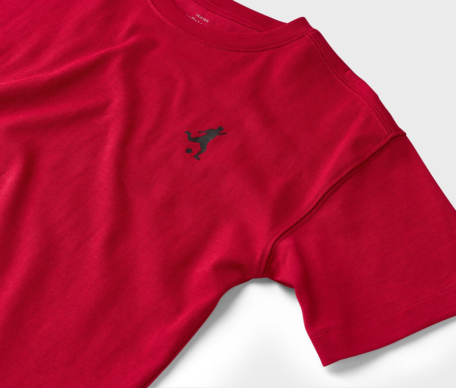 Gros plan d'un t-shirt rouge à manches courtes avec un petit logo sombre représentant un joueur de football.