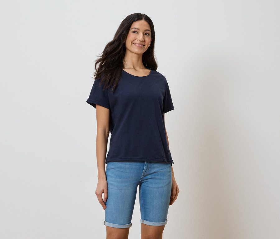 Une femme aux cheveux longs et foncés porte un t-shirt bleu marine et un short en jean bleu sur un fond blanc.