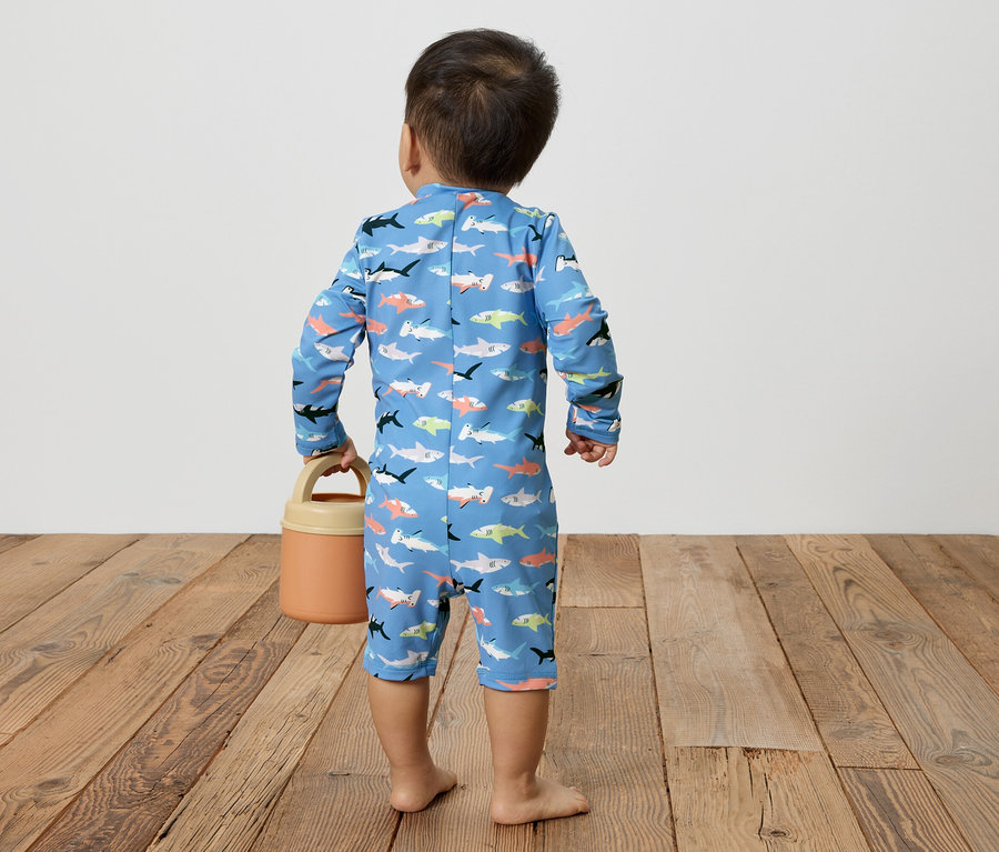 Petit enfant vu de dos portant un maillot de bain bleu à motifs de poissons tenant un arrosoir orange sur un plancher en bois.