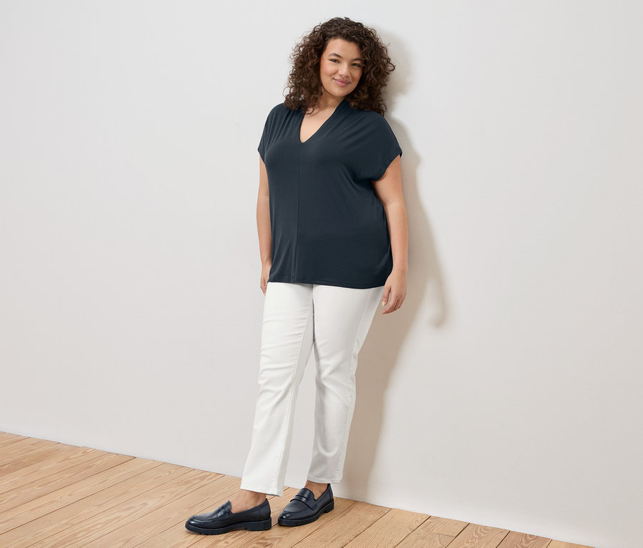 Femme aux cheveux bouclés portant un t-shirt bleu marine, un pantalon blanc et des mocassins bleu marine, appuyée contre un mur blanc.