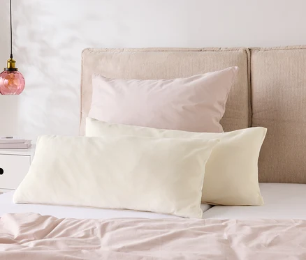 Un lit avec du linge de lit rose clair et deux oreillers crème devant une tête de lit beige. Une lampe est posée sur la table de chevet.