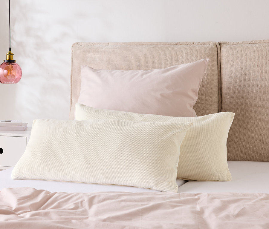 Un lit avec du linge de lit rose clair et deux oreillers crème devant une tête de lit beige. Une lampe est posée sur la table de chevet.