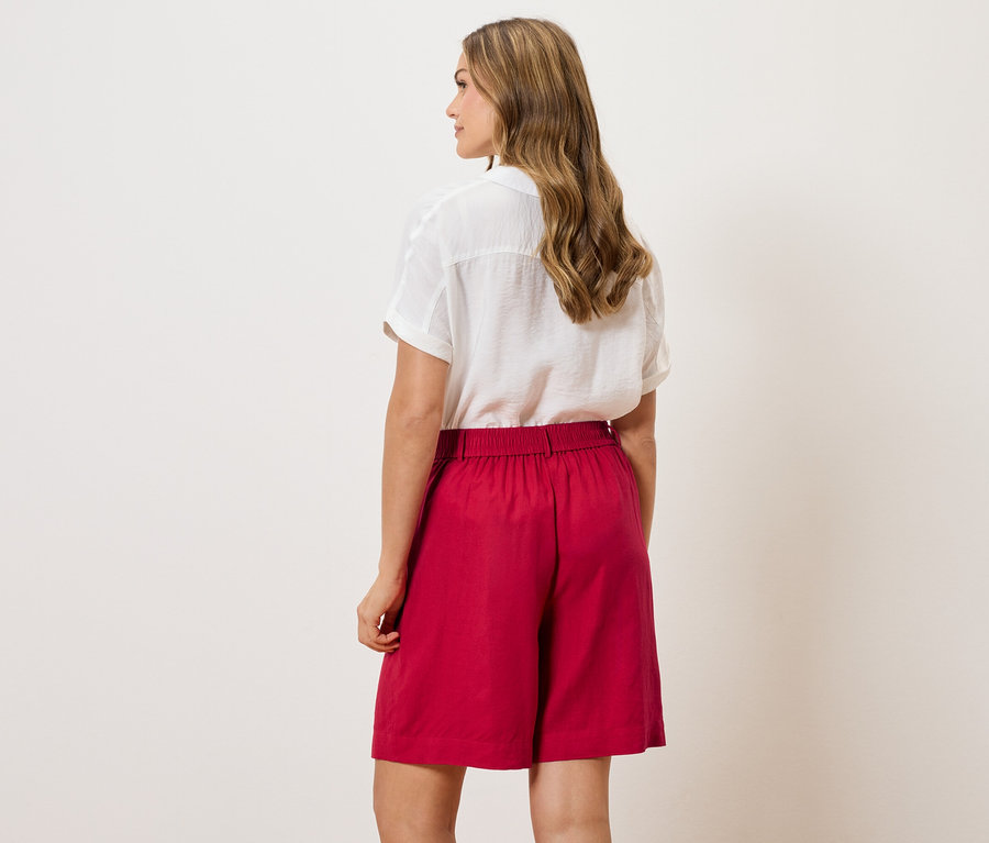 Vue de dos d'une femme aux cheveux ondulés portant un chemisier blanc et un short rouge.
