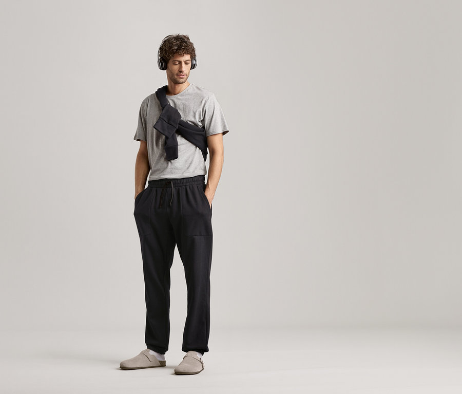 Homme portant un casque audio, un t-shirt gris et un pantalon de survêtement noir sur fond neutre.