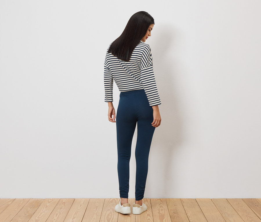 Une femme aux longs cheveux foncés se tient dos à nous, portant un haut rayé, un legging bleu et des chaussures blanches.