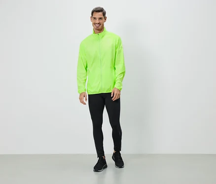 Un homme portant une veste vert néon, des leggings noirs et des baskets se tient devant un mur blanc.
