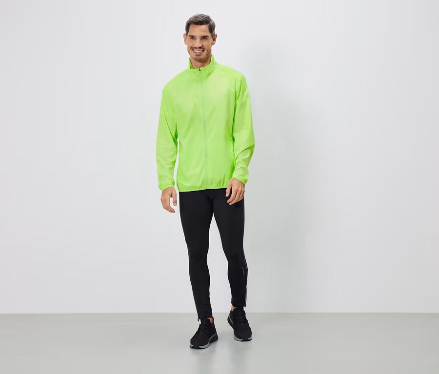 Un homme portant une veste vert néon, des leggings noirs et des baskets se tient devant un mur blanc.